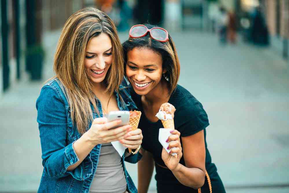 two girls looking at phone and laughing while holding ice cream cones