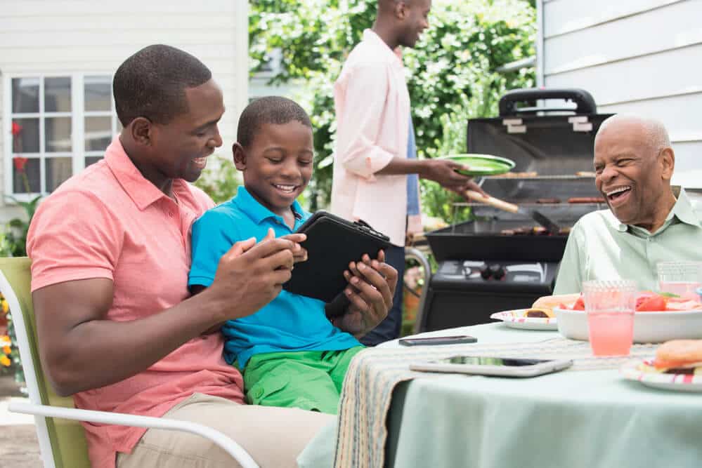 three generation family sitting on patio looking at tablet while cooking out